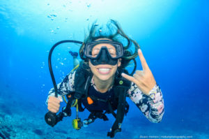 An underwater female diver making the awesome sign and grinning at the camera.